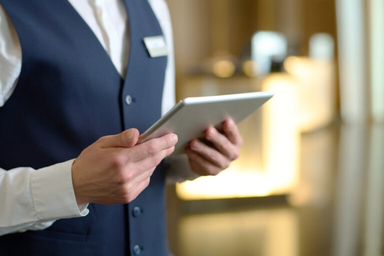 Digital tablet in hands of young male receptionist of luxurious modern hotel standing in lounge by entrance and meeting new guests