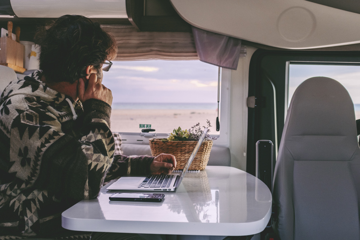 digital nomads Hospitality Trends Post COVID Man working inside camper van sitting at the table and looking outside the window the beautiful beach and ocean in background. Concept of freedom lifestyle and remote online worker people