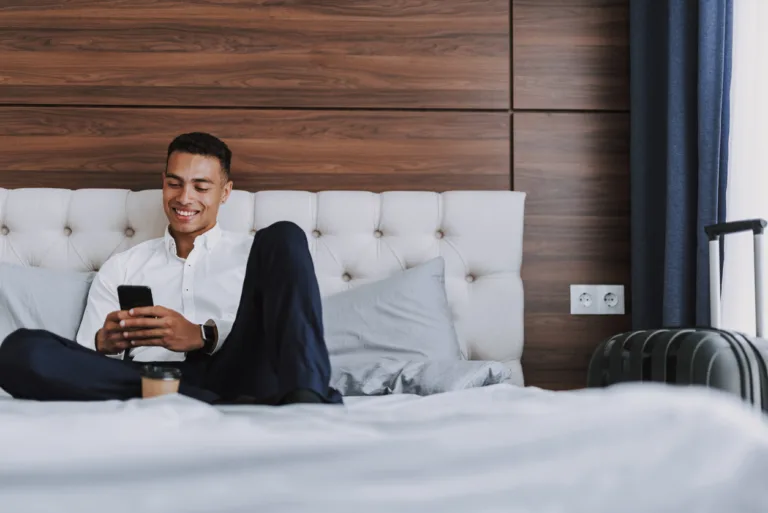 Focus on relaxed young male resting on bed in hotel room. He is having hot drink and using cell phone. Copy space in right side