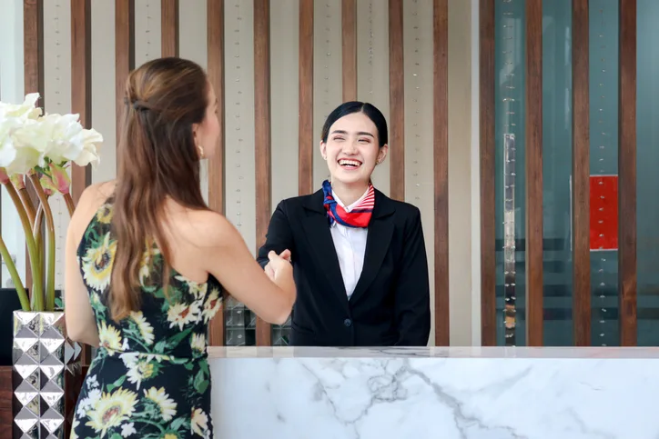 Happy smiling young beautiful Asian female receptionist in suit shaking hands with customer at hotel reception counter desk, check in hotel service on vacation concept.