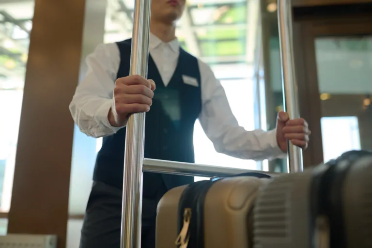 Cropped shot of young bellboy in uniform holding by metallic handles of baggage cart with suitcase of guests of modern hotel