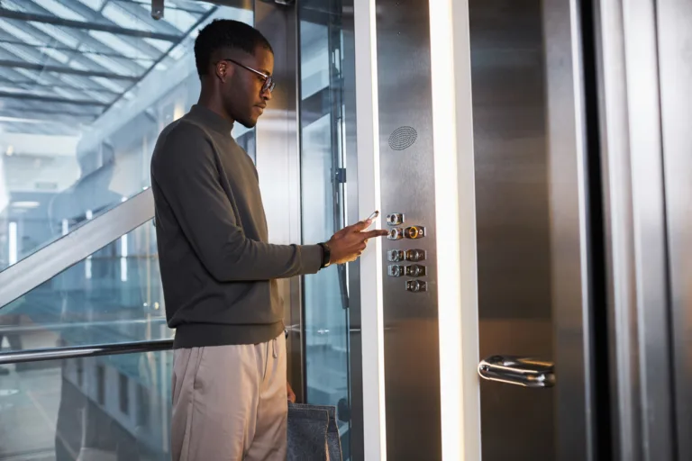 Graphic side view at young black man pushing button in glass elevator in modern office building, copy space
